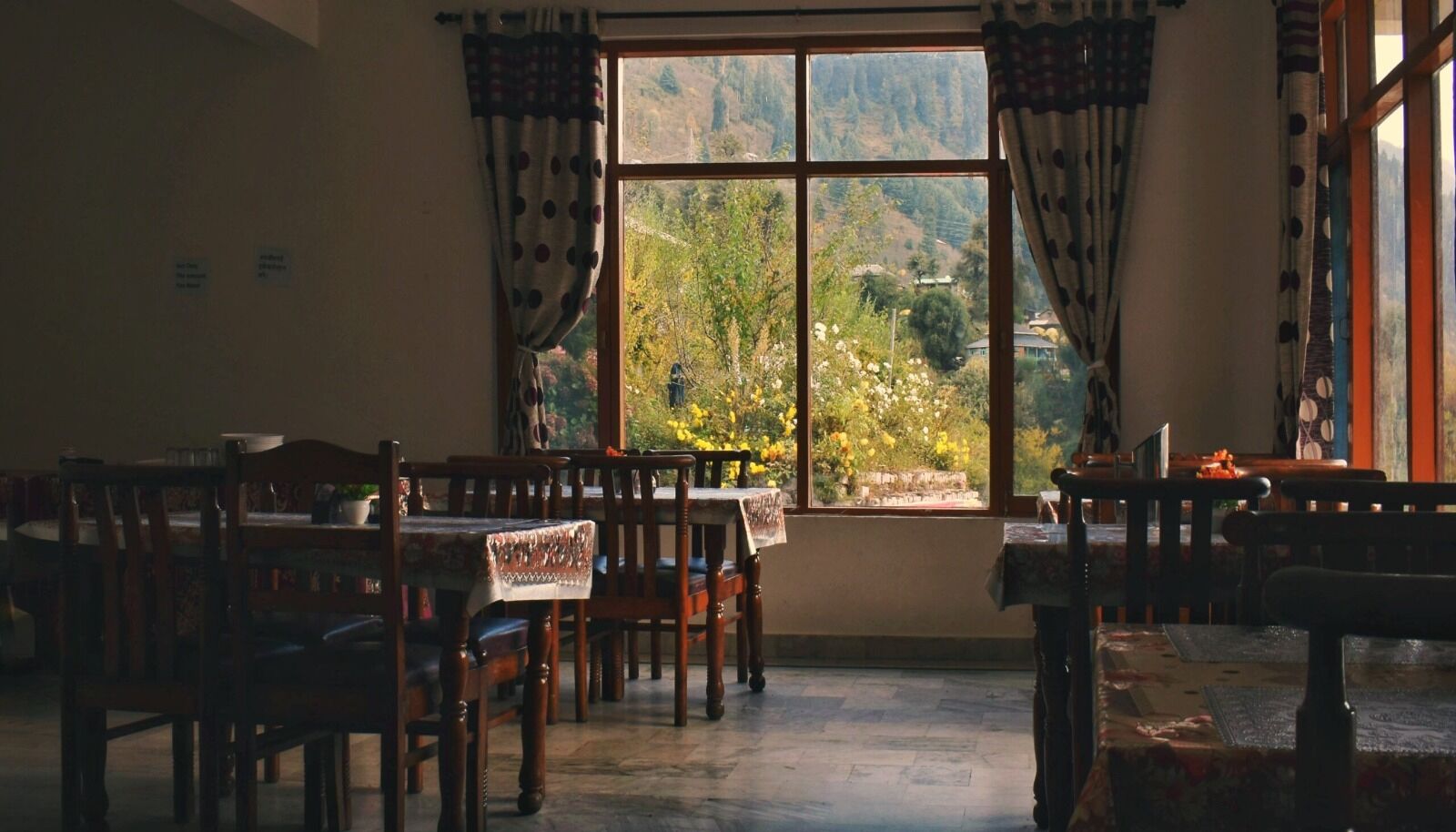 A dining area with wooden tables and chairs, a large window with curtains, and a view of a mountainous landscape.