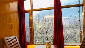 A room with a wooden table, two chairs, a window with red curtains, and a view of buildings and trees outside. - Shreshtam Lakhnpal Resort (Manali)