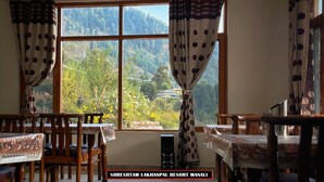 A dining area with wooden chairs and tables, a large window with curtains, and a view of a mountainous landscape. - Shreshtam Lakhnpal Resort (Manali)
