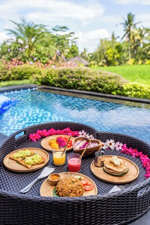 A dining setup by a poolside with a wicker table, plates of food, and a drink.
