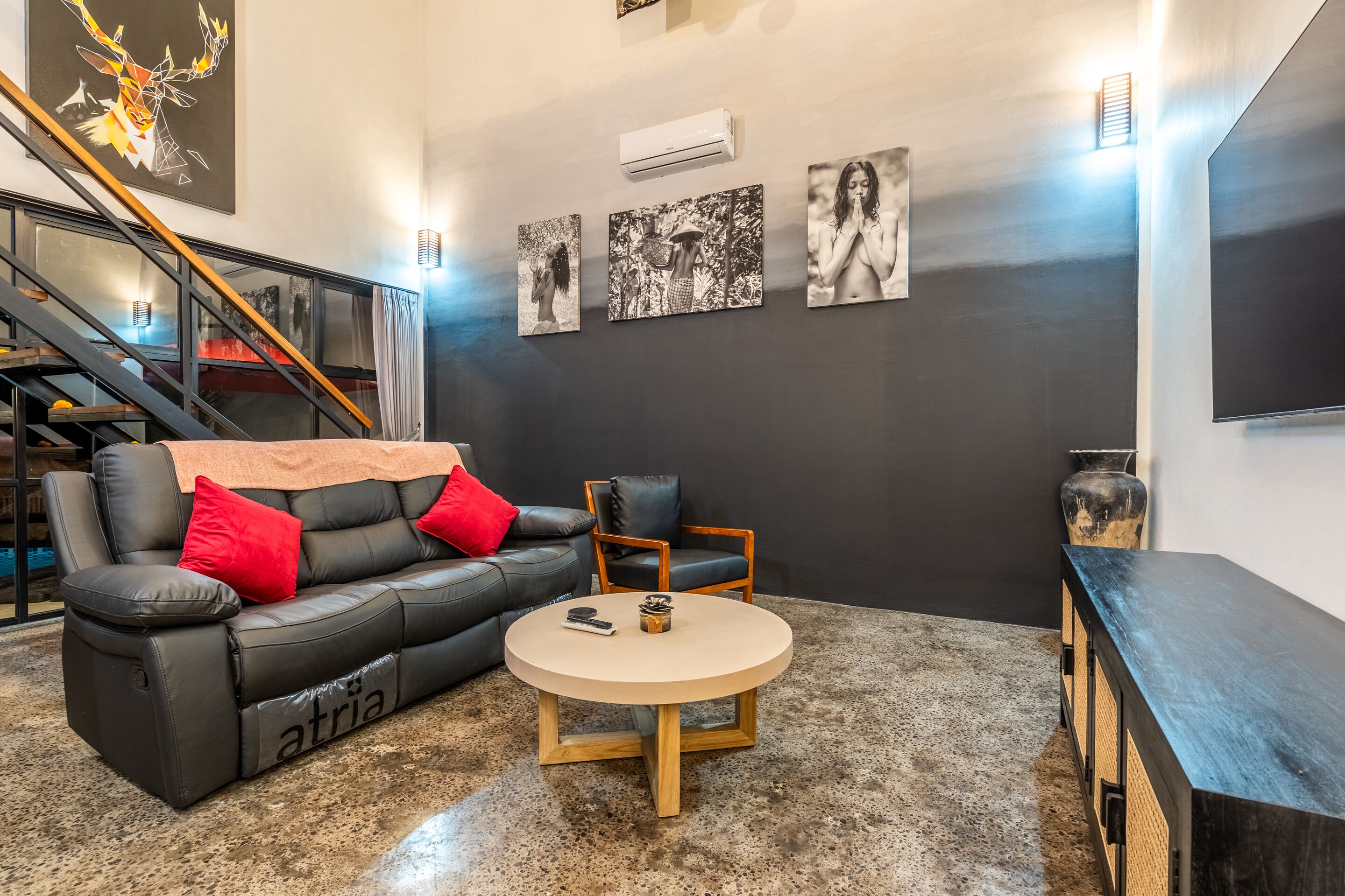 Un salon moderne avec un canapé en cuir, une table basse ronde et un téléviseur. Trois tableaux encadrés ornent le mur, et un escalier se trouve au fond de la pièce.