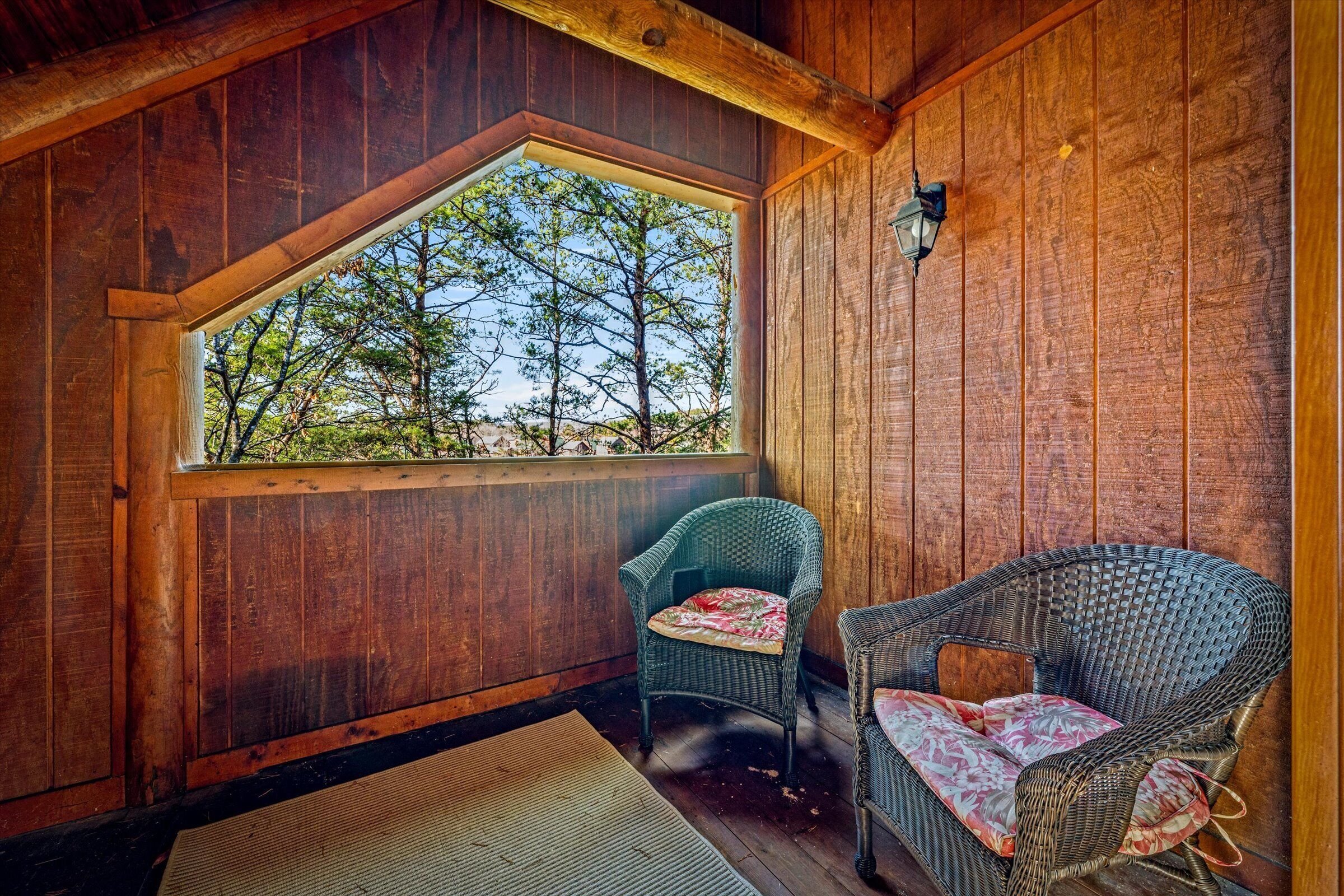 A wooden cabin with a large window overlooking trees, two wicker chairs, and a wooden floor.
