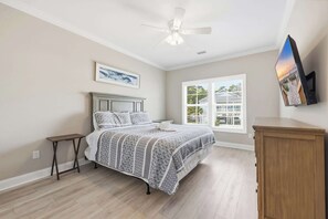 Une chambre à coucher comprenant un lit, une table de chevet, une commode, un ventilateur de plafond, une fenêtre avec vue et un téléviseur fixé au mur.
