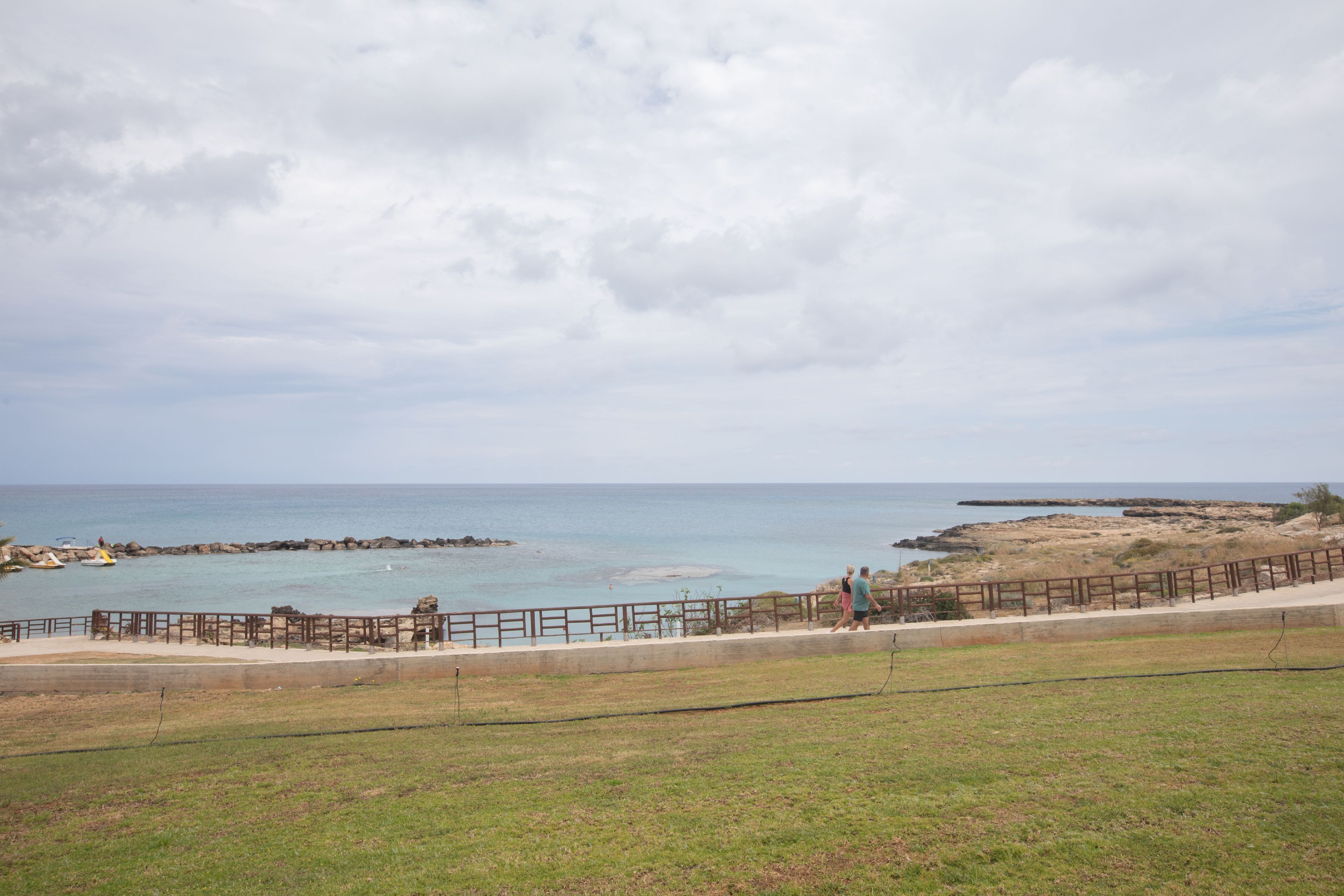 A coastal walkway with a wooden fence, grassy areas, and rocky outcrops.