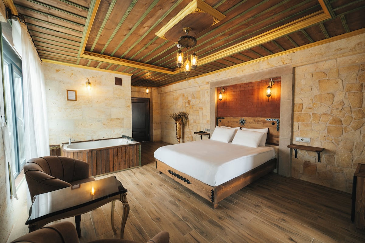 A bedroom with a large bed, wooden floor, stone walls, a wooden ceiling, a desk, a chair, a small table, and a bathroom with a bathtub.