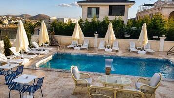 Un área de piscina con sillones de descanso y sombrillas, una mesa con sillas y un edificio al fondo.