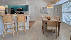Une cuisine moderne avec des meubles de couleur turquoise, un coin repas avec des chaises en osier et une grande fenêtre avec des stores.