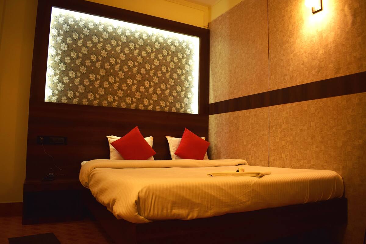 A hotel room with a bed, a nightstand, a window with floral blinds, and a wall-mounted light.