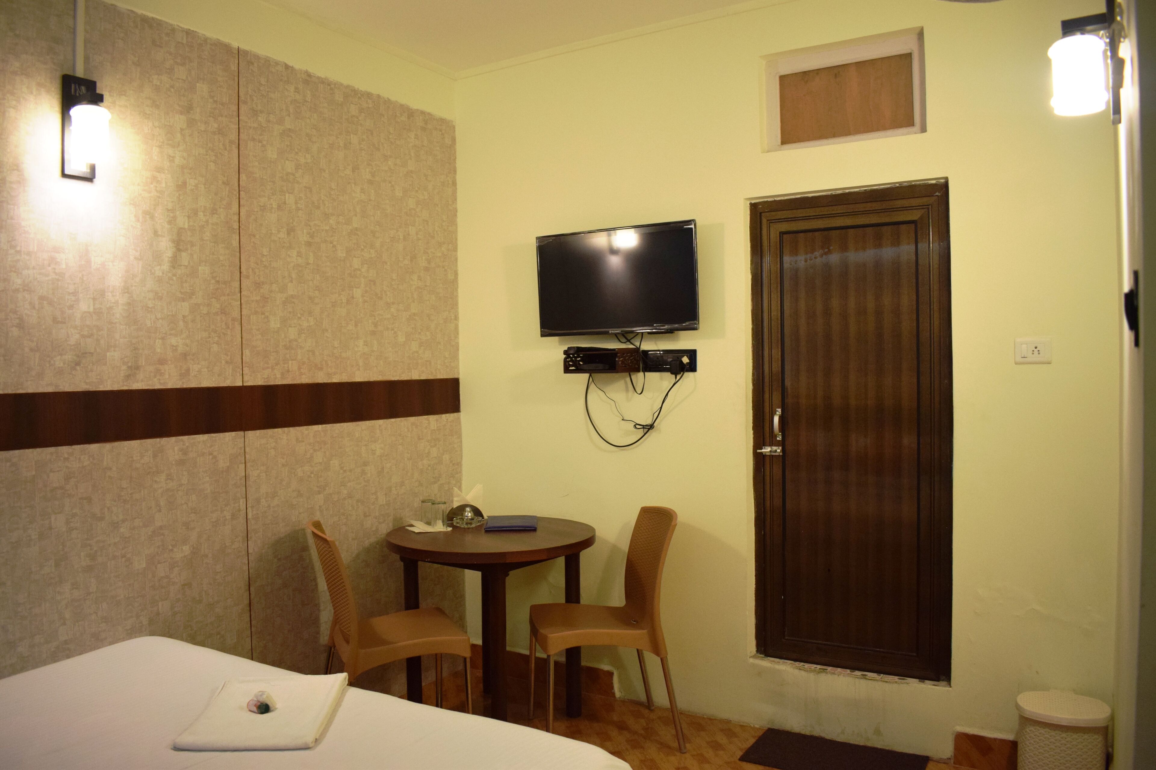 A hotel room with a bed, a small table with two chairs, a television mounted on the wall, and a wooden door.