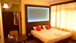 Habitación de hotel con una cama grande, una mesita de noche con lámpara, una pequeña mesa con una silla y un ventanal con cortinas.