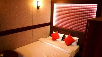 A hotel room with a bed, a nightstand, a red round table, a wall-mounted light, and a window with blinds.