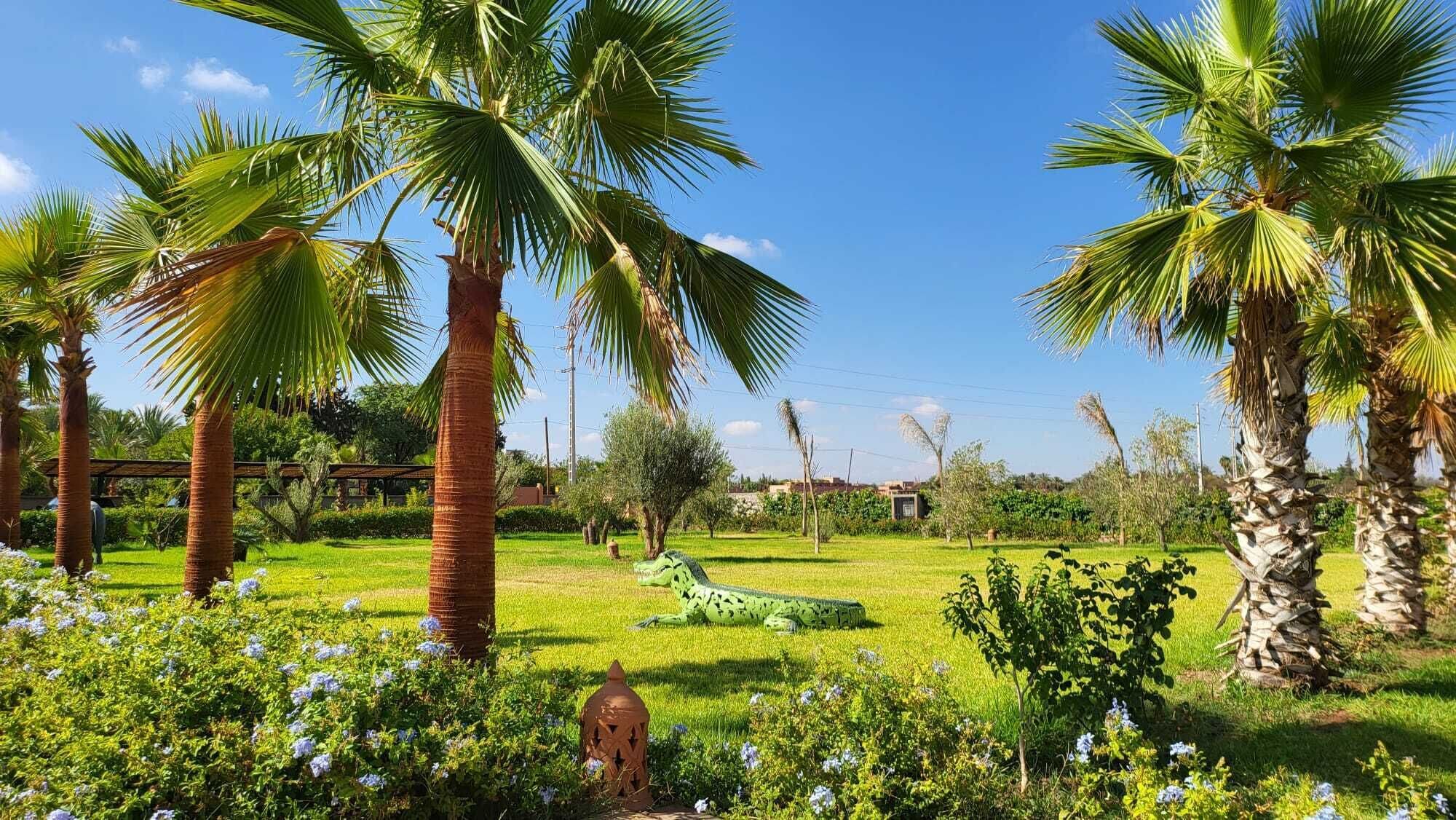 A garden with palm trees, a green sculpture, and a small building in the background.