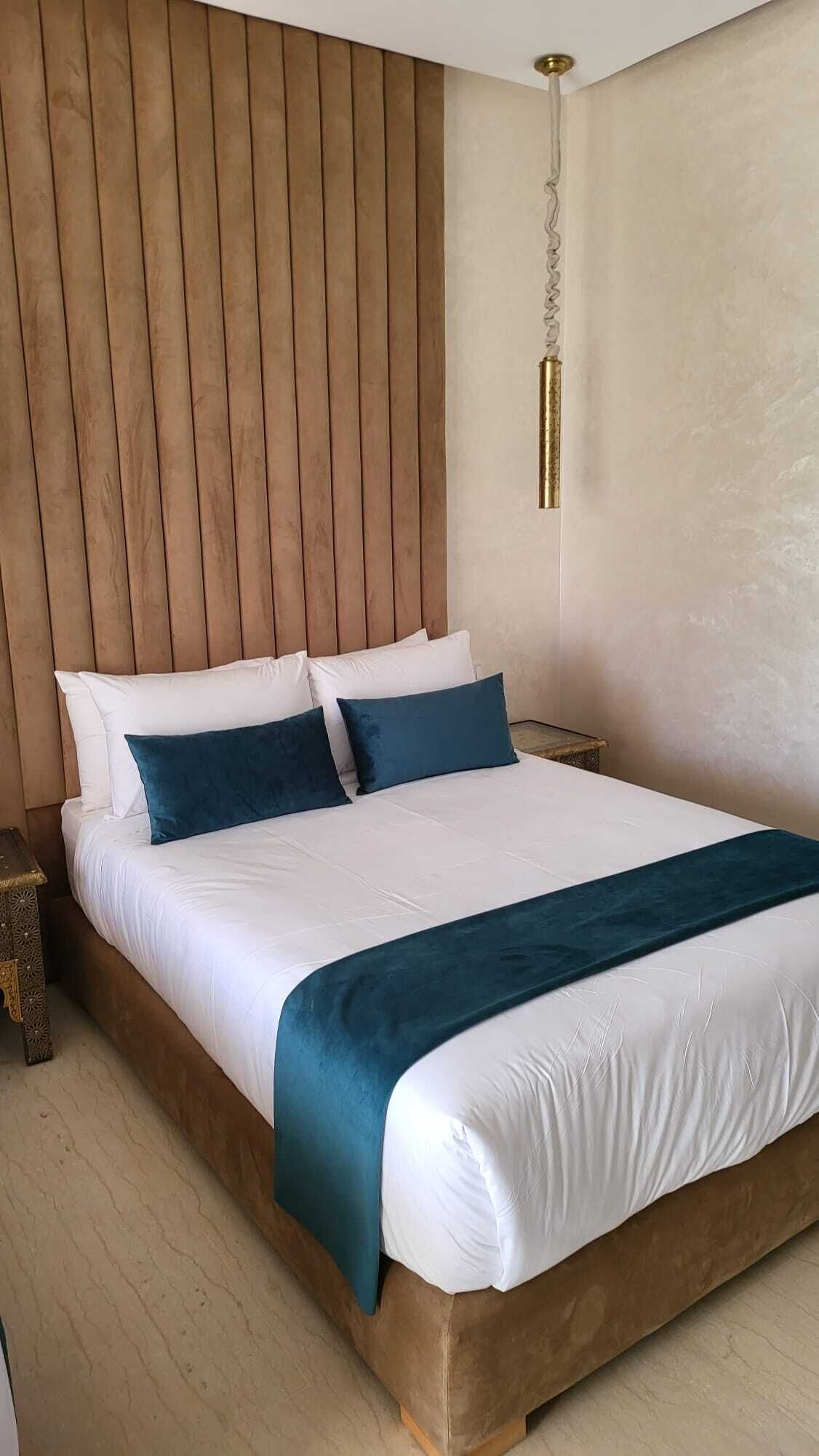 Uma cama grande com roupa de cama branca e verde-água, cabeceira de madeira e luminária pendente.