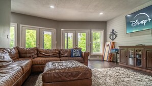 A spacious living room with a large brown leather sectional sofa, a fluffy white rug, and a television mounted on the wall displaying the Disney+ logo. - Lake Effect on Norris Lake (New Tazewell)