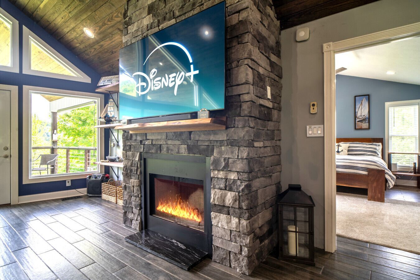 A living room with a stone fireplace, a mounted TV displaying the Disney+ logo, and a view of a bedroom through an open door.