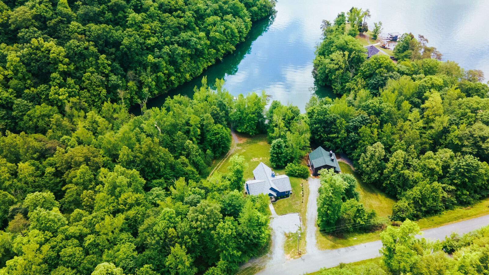 Vue aérienne d’une propriété comprenant deux bâtiments principaux, entourée d’une forêt dense et bordée d’un plan d’eau.
