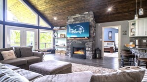 A living room with a stone fireplace, a large flat-screen TV mounted above it, a cozy seating area with a sofa and ottomans, and a kitchen area with bar stools. - Lake Effect on Norris Lake (New Tazewell)