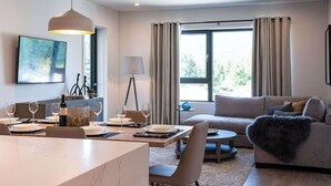 A modern living room with a dining area, a sofa, a coffee table, and a large window with curtains. - The Stoke Stopover - Revelstoke Condo with Hot Tub (Revelstoke)
