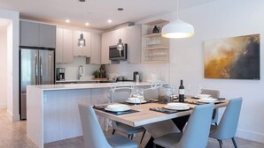 A modern kitchen with a central island, stainless steel appliances, and a dining area with a table set for four. - The Stoke Stopover - Revelstoke Condo with Hot Tub (Revelstoke)