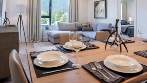 A dining area with a table set for two, a sofa in the background, and a view of greenery outside. - The Stoke Stopover - Revelstoke Condo with Hot Tub (Revelstoke)