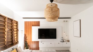 A modern living room with a large flat-screen TV mounted on a white wall, a grey sectional sofa, a wicker pendant light, and a wooden slatted window blind. - Casa Don Toribio: Luxury Home with Private Pool (Aguada)
