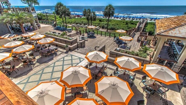 An outdoor patio area with multiple tables and chairs, surrounded by orange umbrellas, overlooking a beach and ocean.