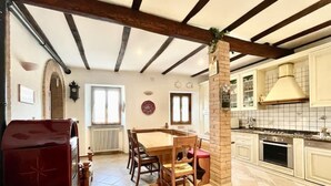 A dining area with a wooden table and chairs, a red upholstered chair, a brick pillar, and a kitchen with white cabinets and a stove. - Mazzetti Country House-Country living and garden (Collina di Savignano)