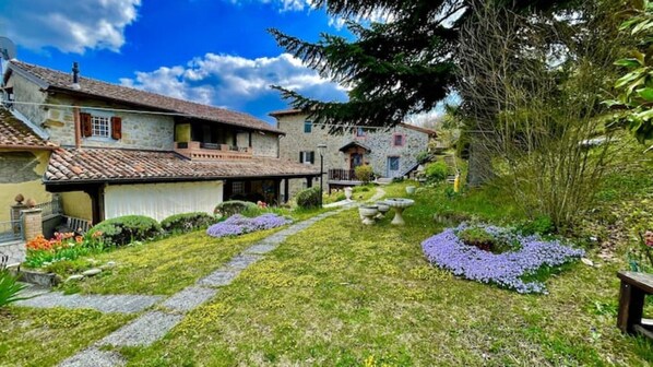 A stone building with a tiled roof, a stone pathway, and a garden with purple flowers.