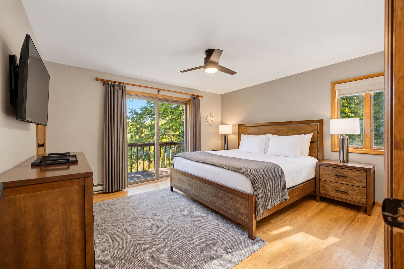 A bedroom with a bed, a TV, a desk, a nightstand, a lamp, a ceiling fan, and a view of trees through a window.