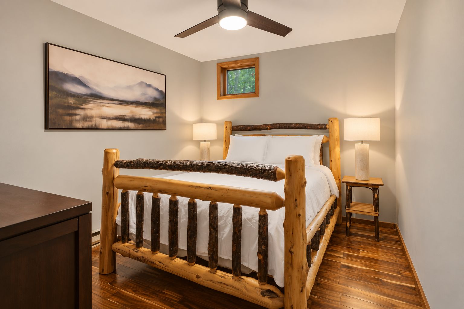 A room with a wooden bed, two bedside lamps, a ceiling fan, a framed picture on the wall, and a window with a view of trees.