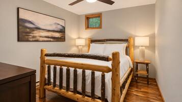 A room with a wooden bed, two bedside lamps, a ceiling fan, a framed picture on the wall, and a window with a view of trees.