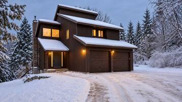 A two-story wooden house with a garage, surrounded by snow-covered trees.