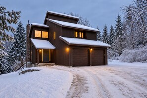 A two-story wooden house with a garage, surrounded by snow-covered trees.