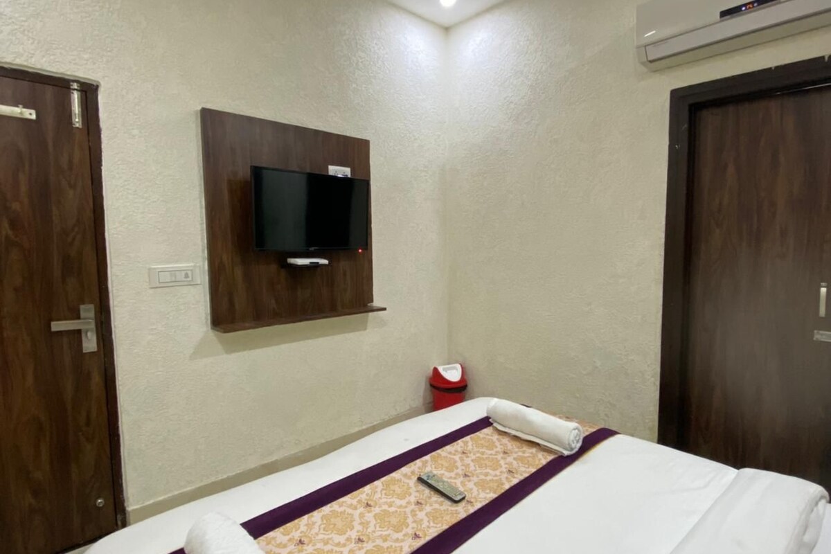 A hotel room with a bed, a TV mounted on the wall, a red trash bin, and a wooden door.