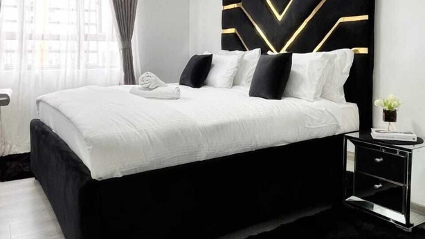 A modern bedroom with a black and white bed, a geometric headboard, and a small bedside table with a book and flowers. - Lux Suites Gold Park Apartments Kilimani (Nairobi)