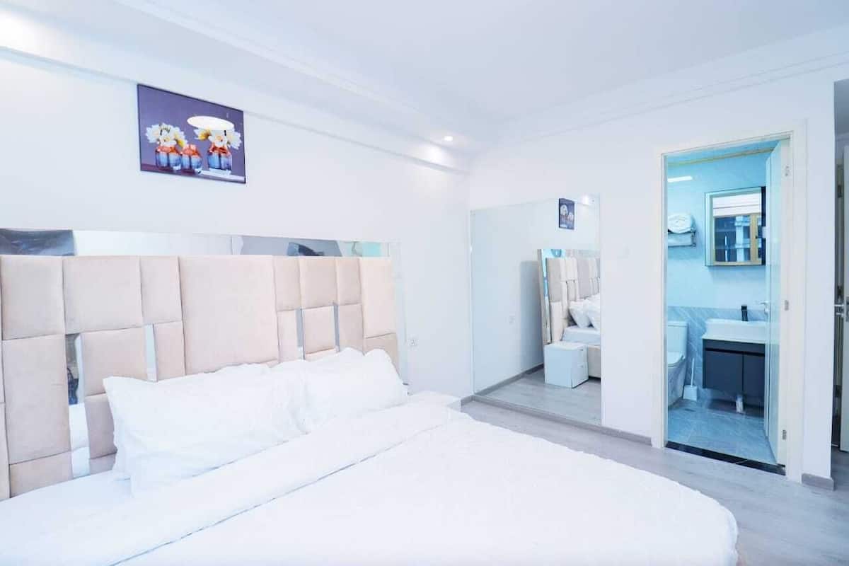 Una habitación de hotel moderna con una cama grande, una mesita de noche, un espejo y un baño visible a través de una puerta abierta.