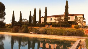 Un espace piscine paisible, où se reflètent de grands cyprès et où l’on aperçoit un bâtiment doté d’un toit en tuiles.