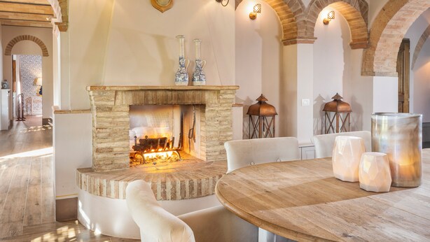 Un coin chaleureux avec une cheminée en pierre, deux appliques murales et une décoration représentant une tête de cerf. On y trouve deux chaises blanches et une table ronde en bois sur laquelle sont posés trois chandeliers aux formes géométriques.
