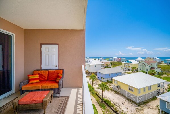 Un balcón con sofá y reposapiés de mimbre, una puerta y vistas a casas de playa.