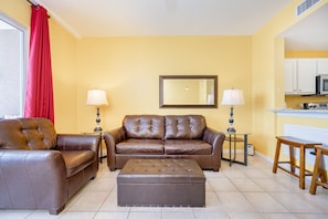 A living room with a brown leather sofa, a matching ottoman, two side tables with lamps, a red curtain, and a kitchen area in the background. - The Dunes 503 1 Bedroom Condo (Gulf Shores)
