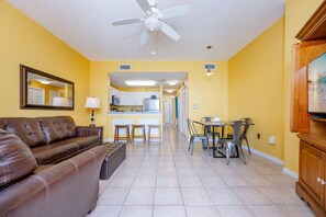A living room with a brown leather sofa, a dining area with a table and chairs, a kitchen with a refrigerator and microwave, and a ceiling fan. - The Dunes 503 1 Bedroom Condo (Gulf Shores)