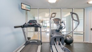 A gym with treadmills and an elliptical machine, a large window, and a flat-screen TV mounted on the wall. - The Dunes 503 1 Bedroom Condo (Gulf Shores)