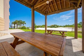 A wooden picnic table under a covered area with a view of a beach and palm trees. - The Dunes 503 1 Bedroom Condo (Gulf Shores)