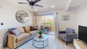 A modern living room with a sofa, armchair, coffee table, and a TV. There's a ceiling fan, a large window with curtains, and a sign that says "RELAX AND UNWIND". - Villa Sunrise 1 Bedroom Condo by RedAwning (Scottsdale)