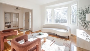 A spacious living room with a white sofa, two pink armchairs, a coffee table, and large windows offering a view of trees. - Private, Serene Mt Lookout 5B Retreat w/Garage & EV Charger (Cincinnati)
