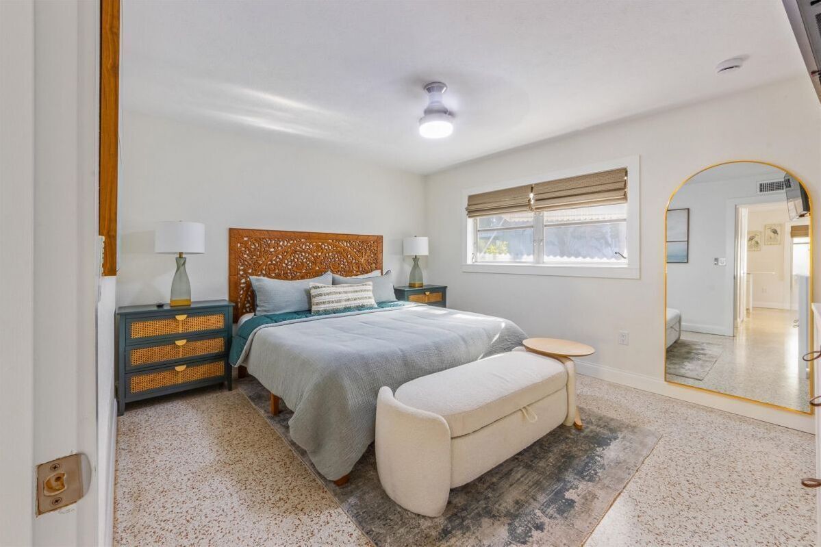 A bedroom with a bed, bedside tables, a bench, a mirror, and a window with blinds.