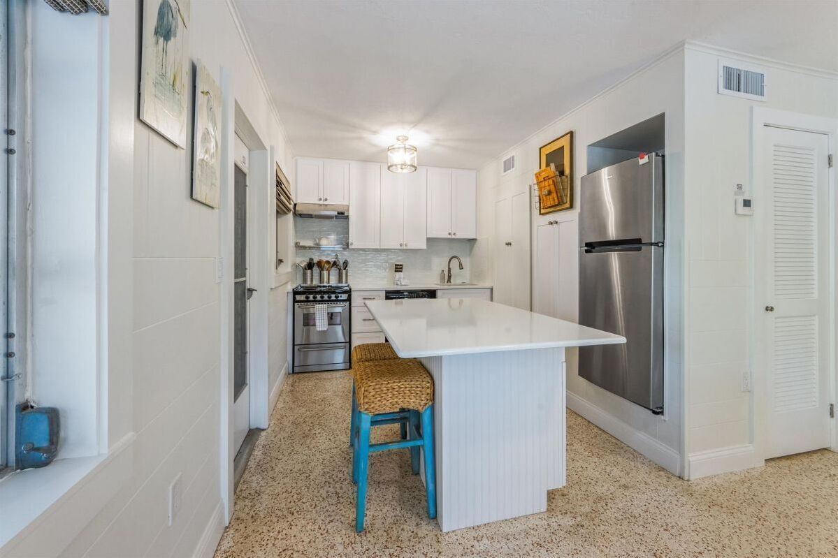 A kitchen with white cabinets, a stainless steel refrigerator, a built-in oven, and a small island with a woven stool.