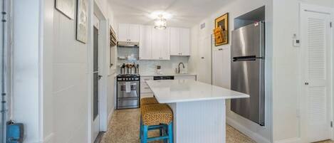 A kitchen with white cabinets, a stainless steel refrigerator, a built-in oven, and a small island with a woven stool.