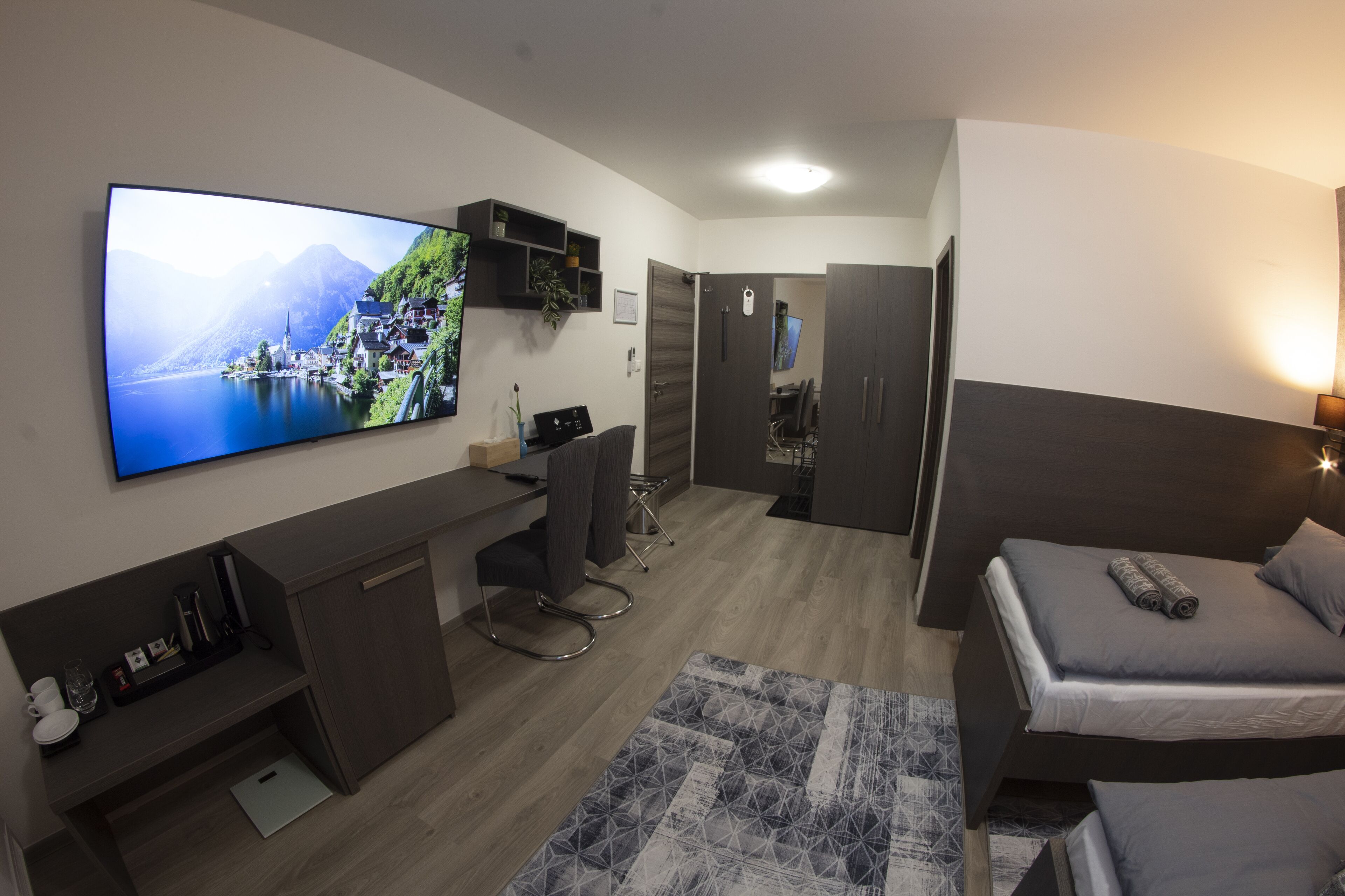 Una habitación de hotel moderna con una televisión de pantalla plana grande, un escritorio con una silla, una cama con almohadas y una pequeña alfombra.