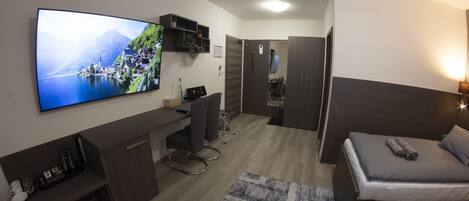 Una habitación de hotel moderna con una televisión de pantalla plana grande, un escritorio con una silla, una cama con almohadas y una pequeña alfombra.